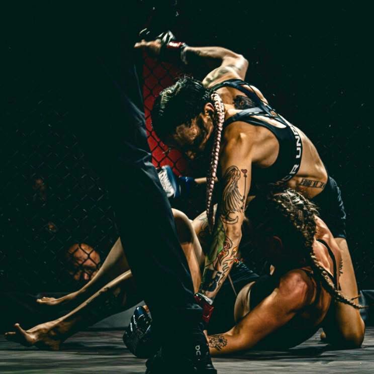 Two women and a man engaged in a mixed martial arts fight inside a cage, with one woman on the ground and the man on top.