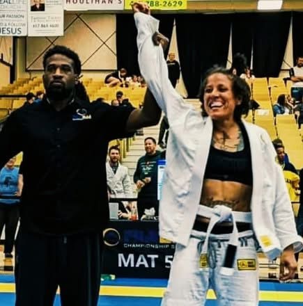 A smiling female athlete in a judo gi holding a trophy, with a man beside her in a sports arena.