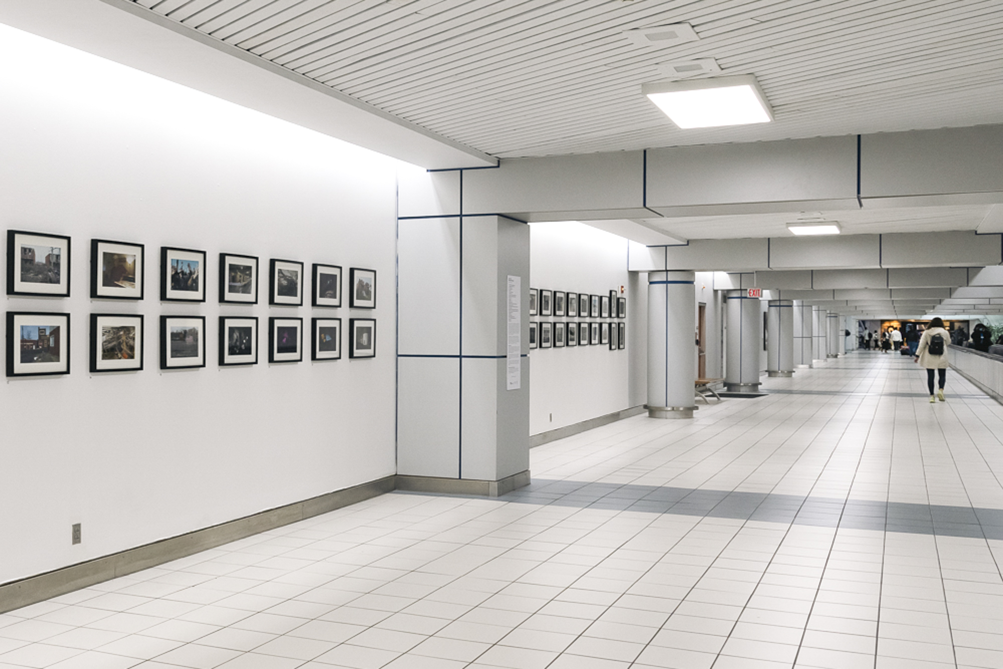Gallery with framed photographs on white wall, tiled floor, and people walking in the distance.