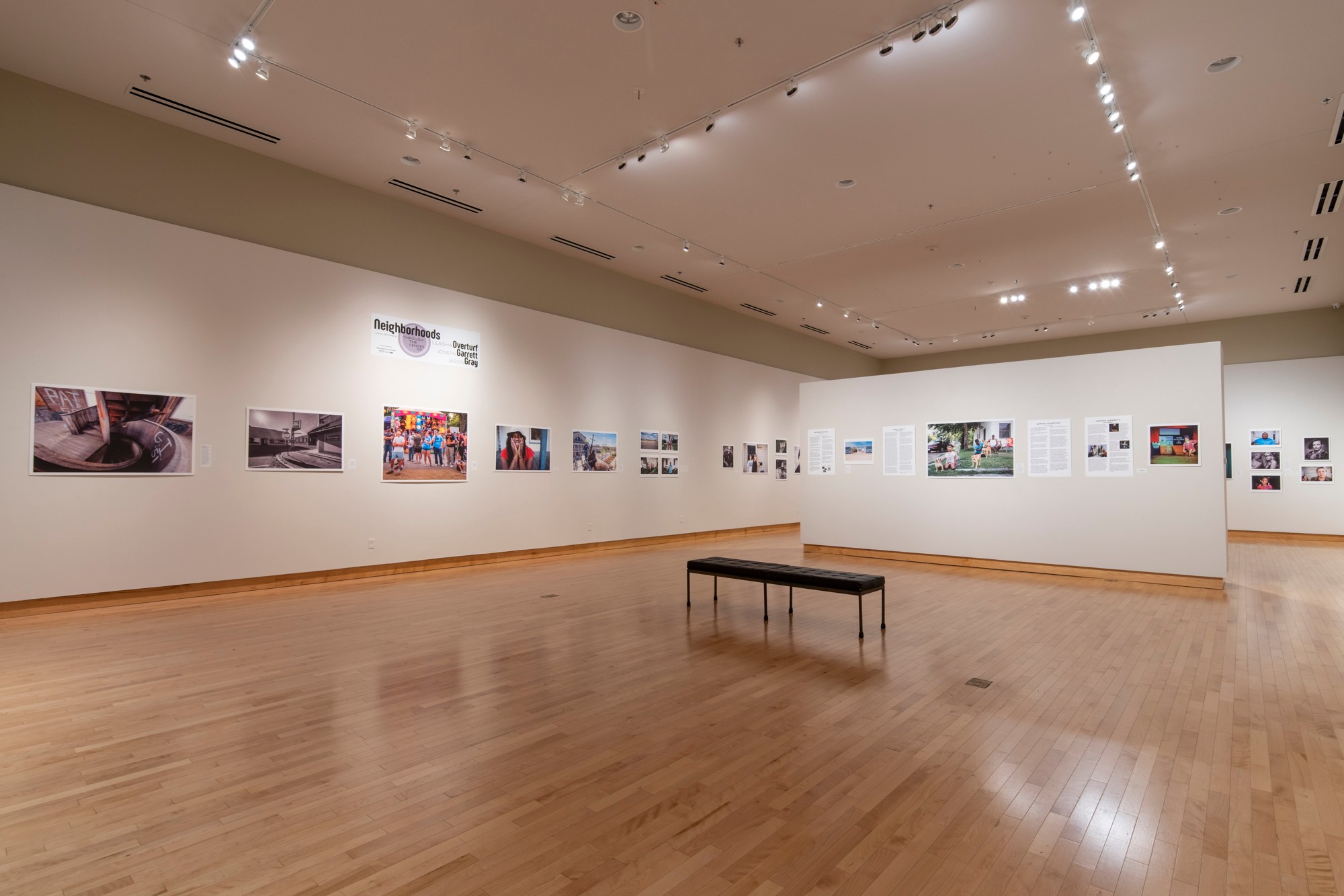 Gallery with white walls displaying framed photographs, wooden floor, and a bench in the center, well-lit ceiling, and no visible visitors.