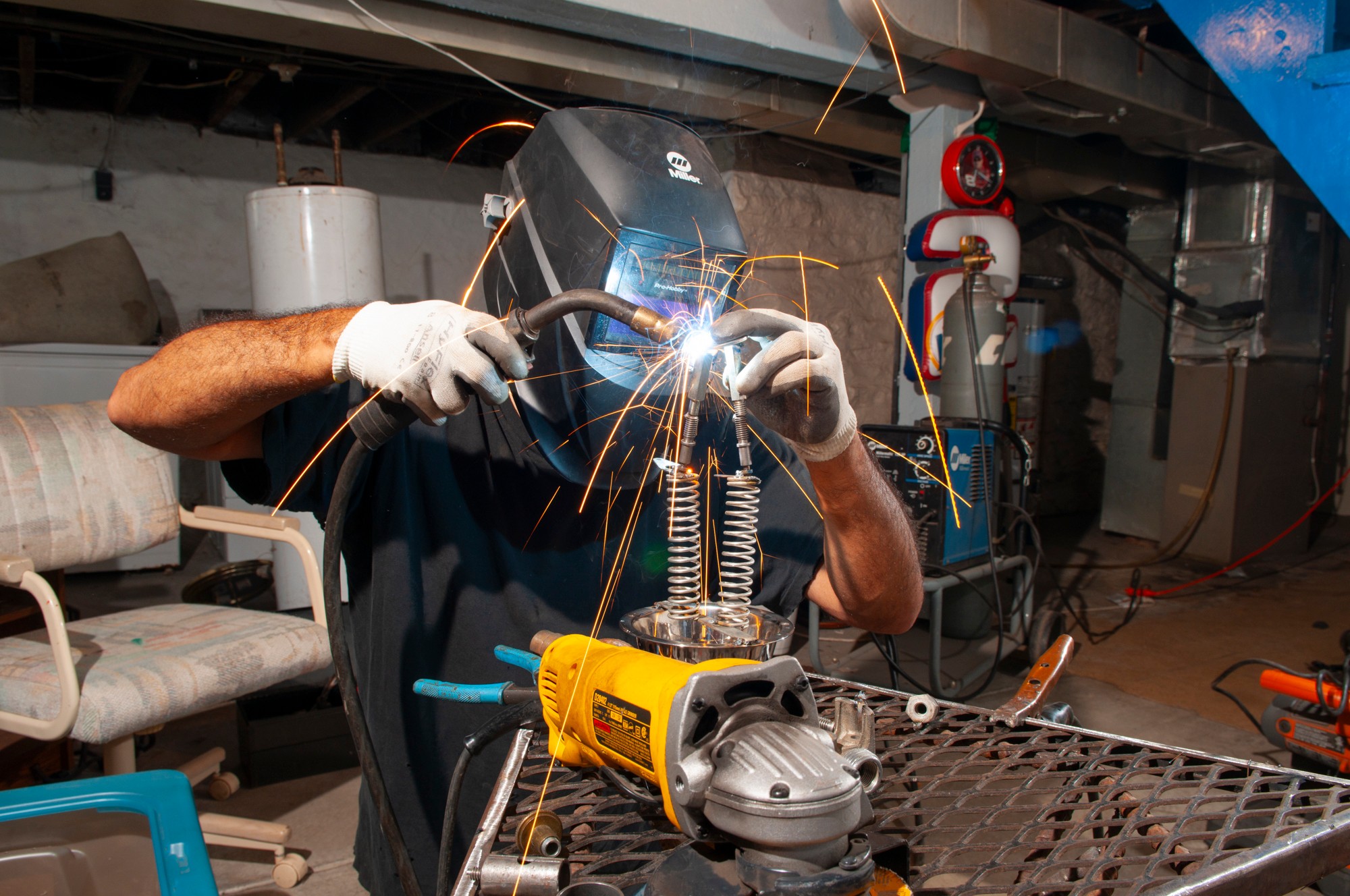 Person welding metal parts with sparks flying, wearing protective gear in workshop, using a yellow power tool on metal surface.