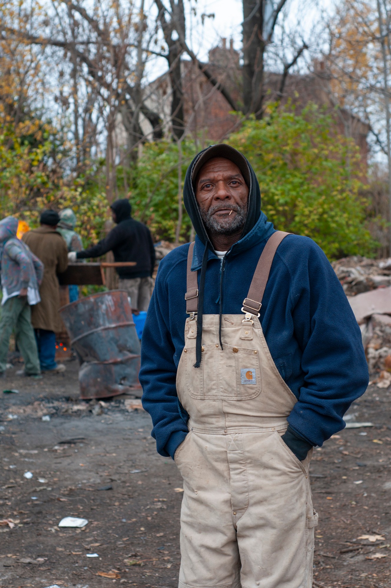 Man in beige overalls and dark hoodie standing outdoors with trees and people in the background.