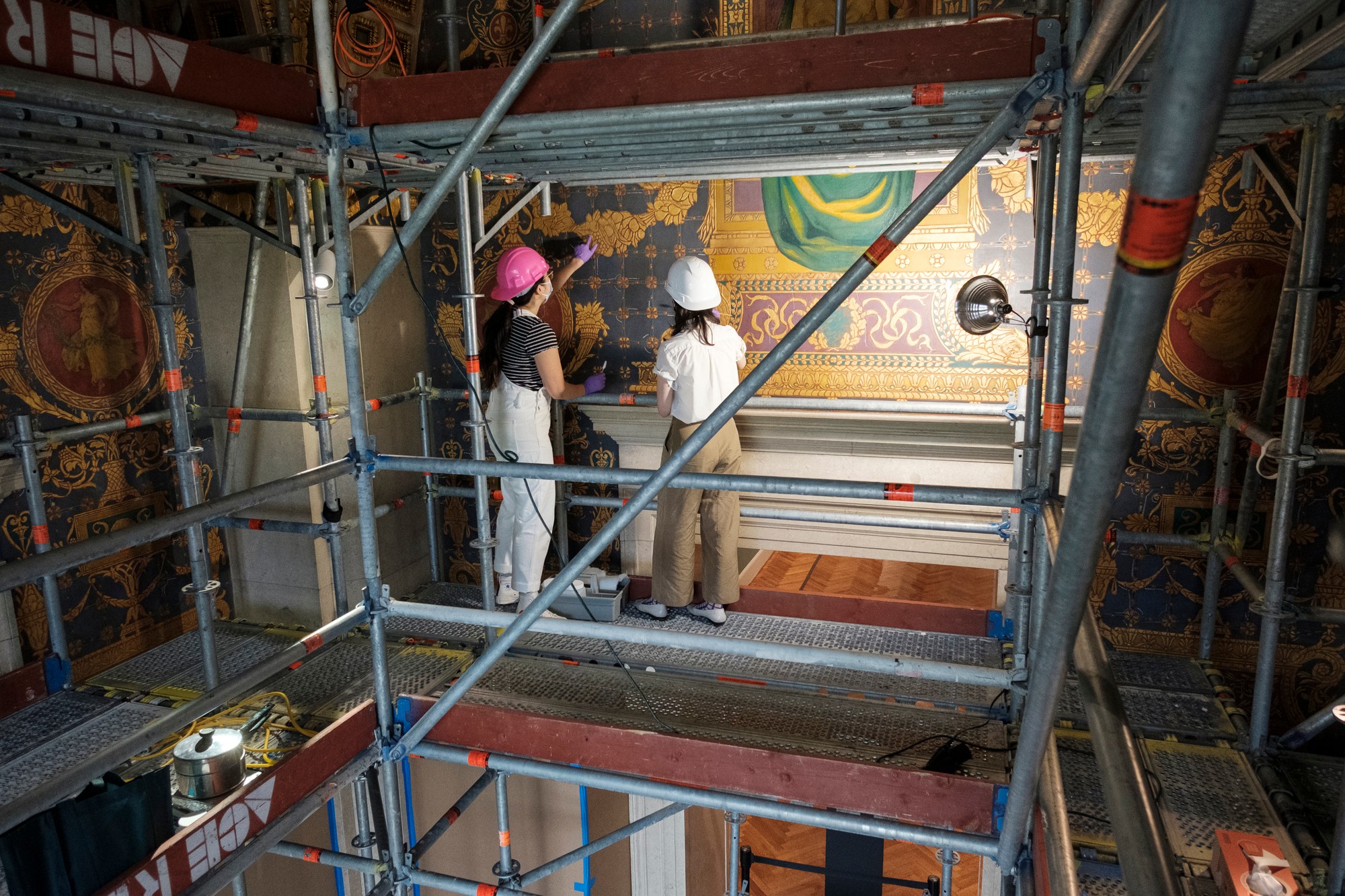 Three people wearing hard hats and safety gear stand on scaffolding, examining a wall with colorful artwork or murals.