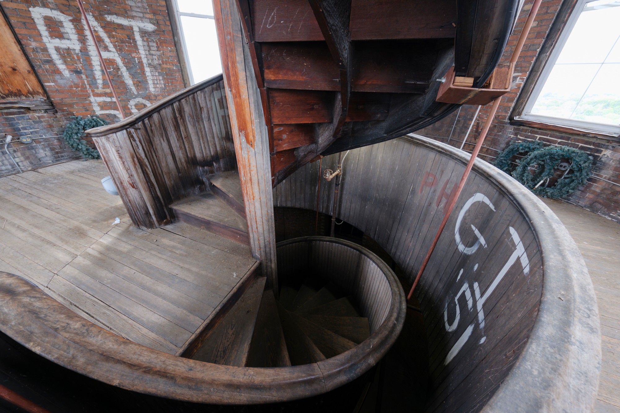 Spiral staircase viewed from above, with wooden steps and metal railing, inside a building with brick walls and windows.