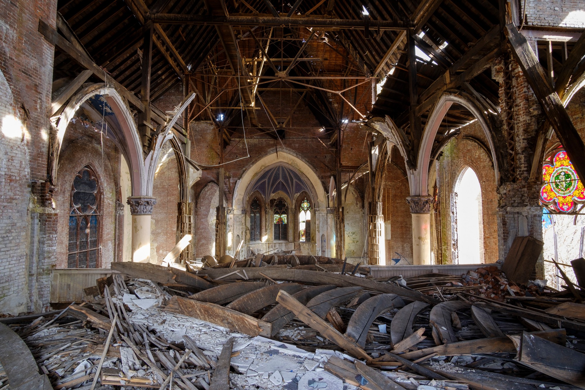 Interior of a damaged church with debris, broken pews, and damaged walls and windows, showing a partially collapsed ceiling.