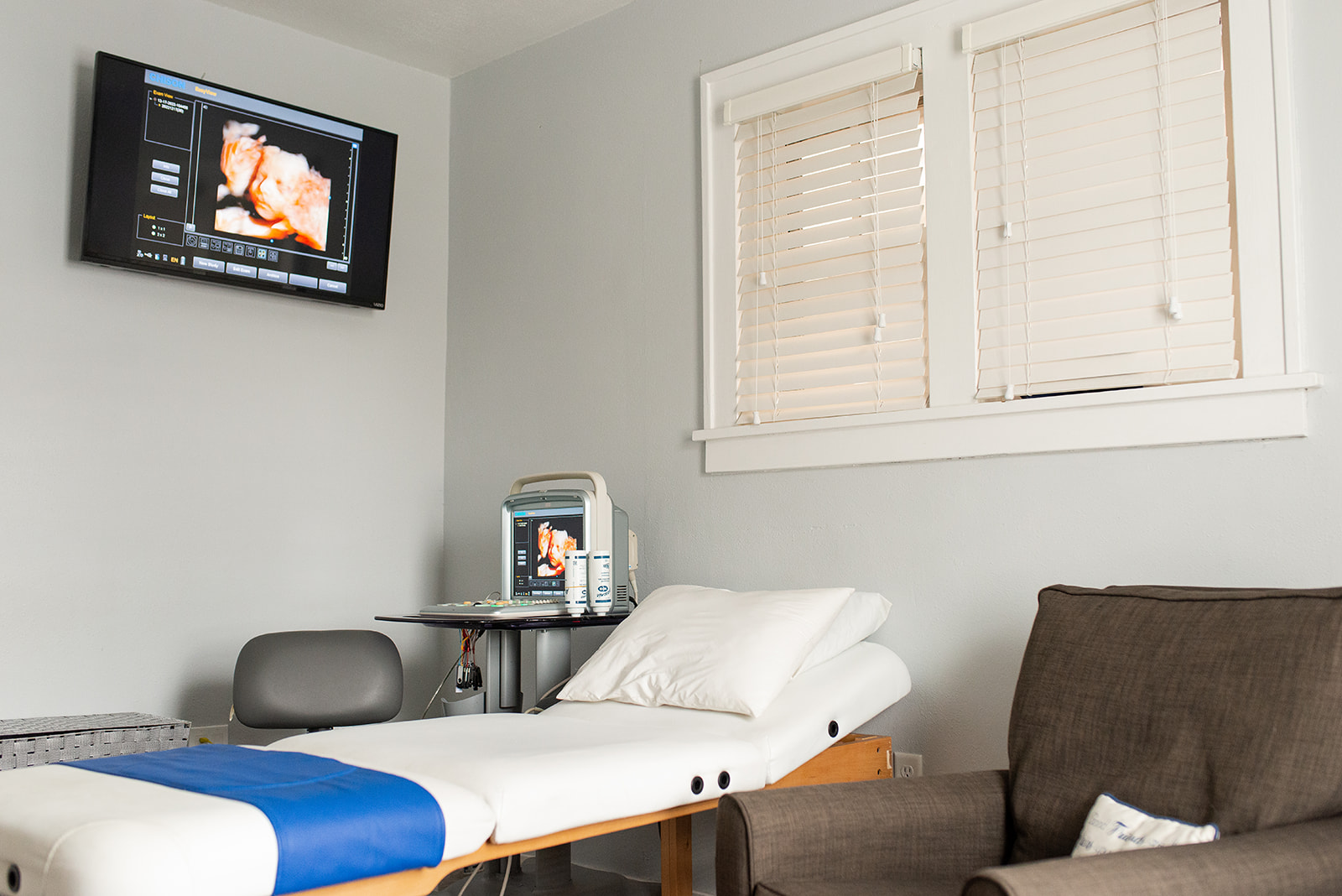 Hospital room with bed, monitor, window with blinds, and brown armchair, all in a light-colored room.