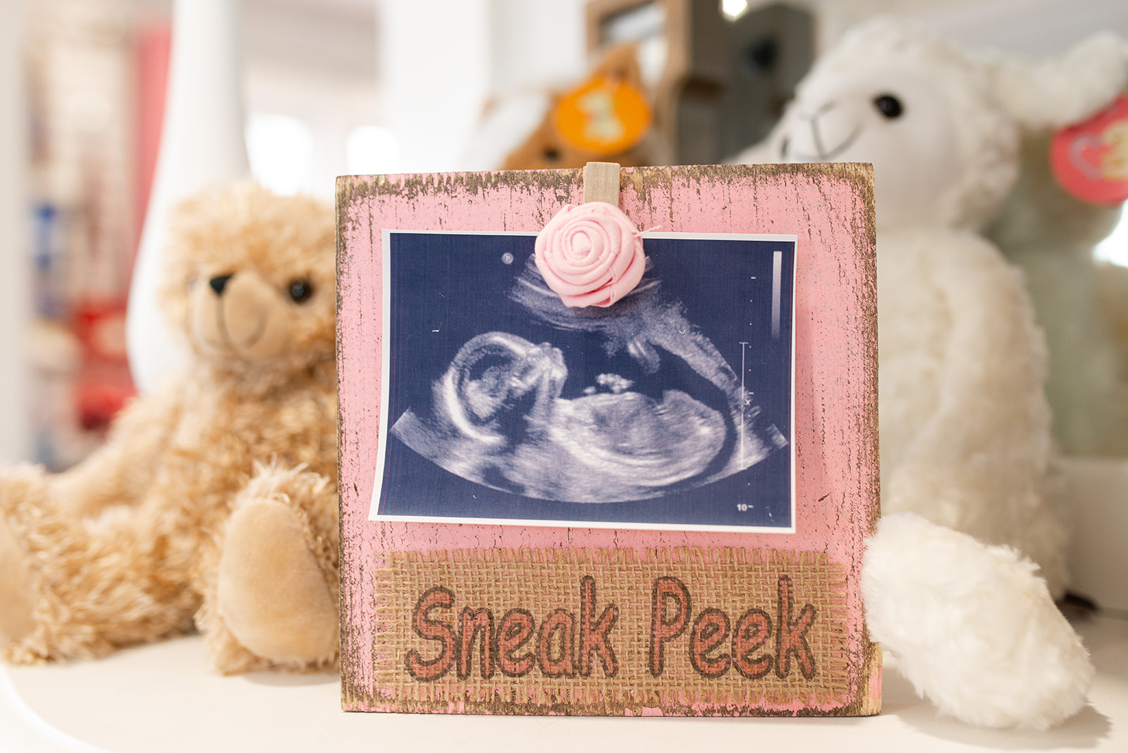 A pink wooden frame with a photo of a fetus ultrasound and the words 'Sneak Peek' below, surrounded by plush toys.