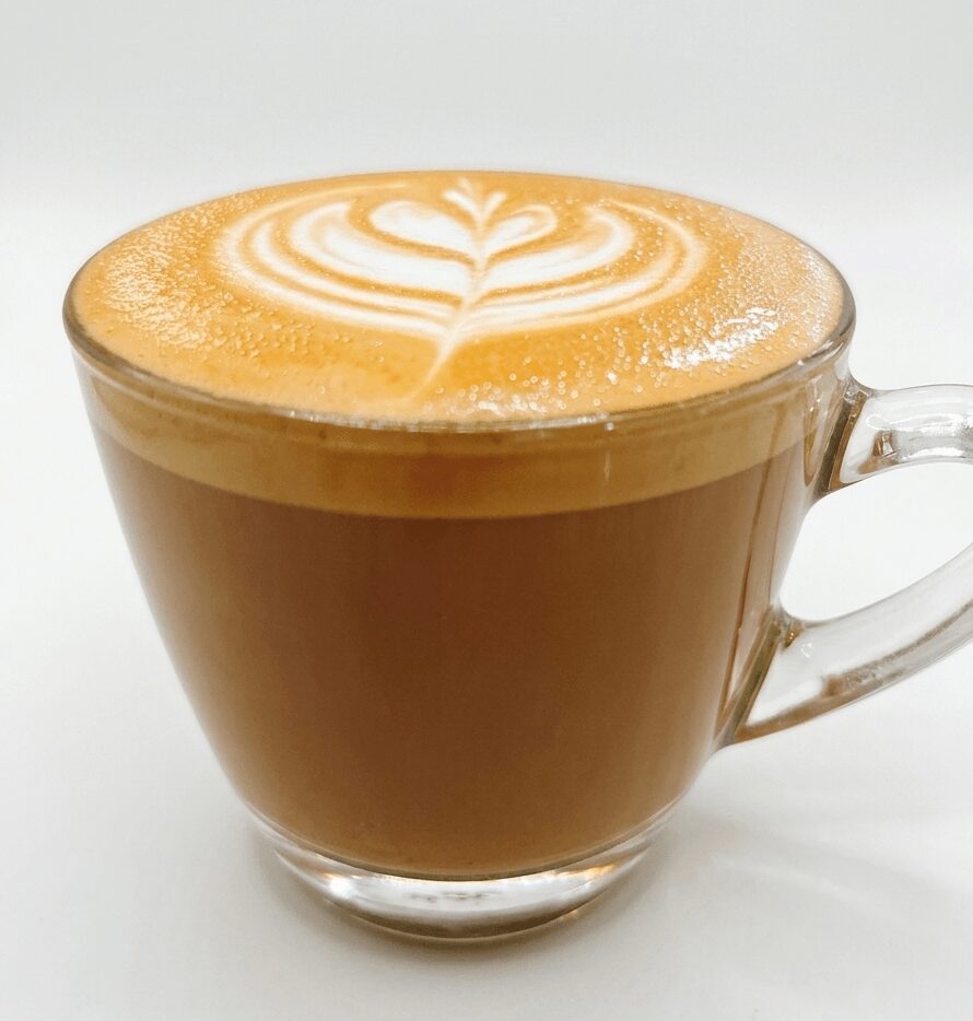 A glass cup filled with coffee topped with latte art in a heart shape, on a white background.