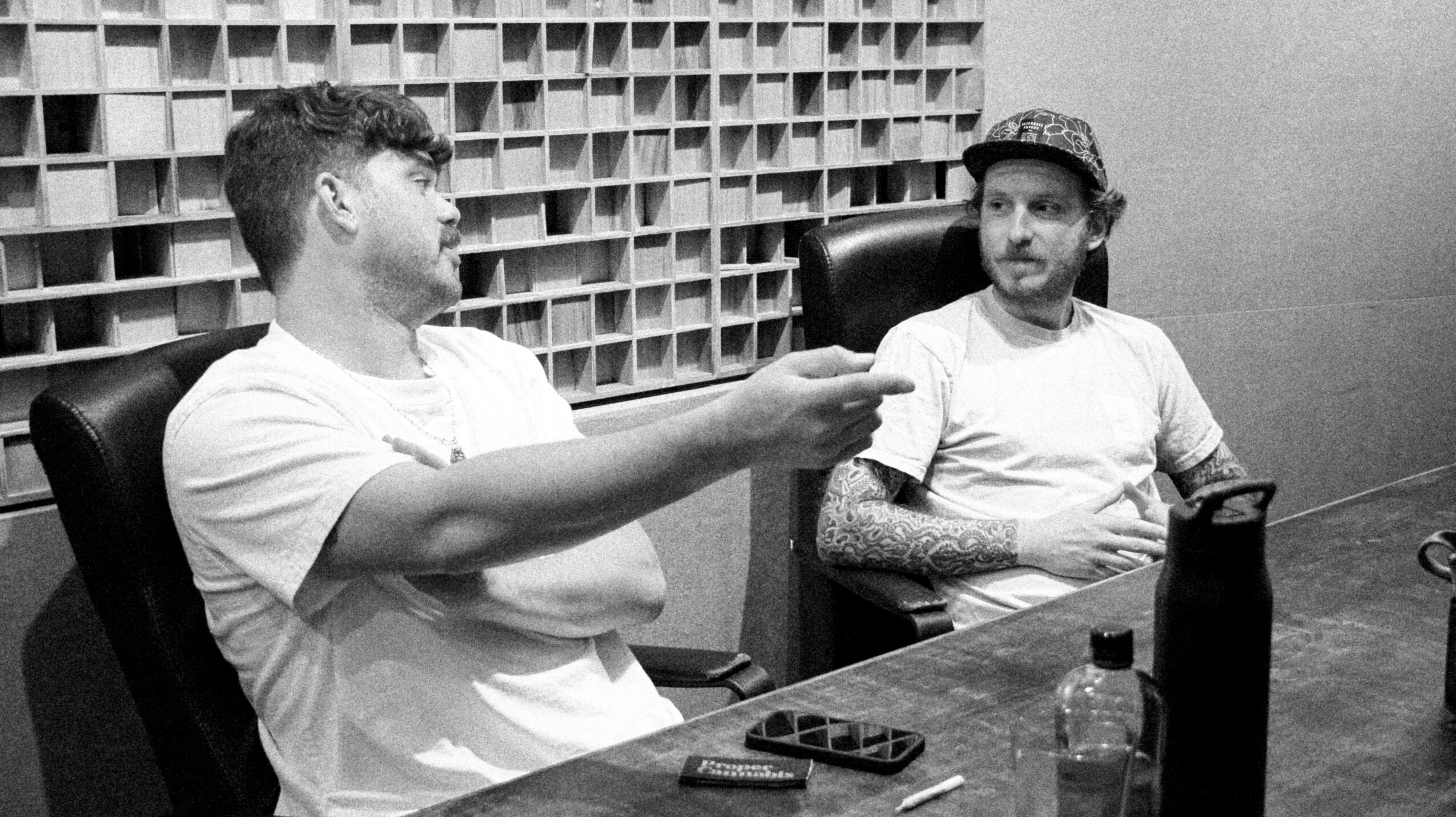 Two men sit at a table in a room with a wall of shelves behind them. One man is smiling and pointing, the other listens.