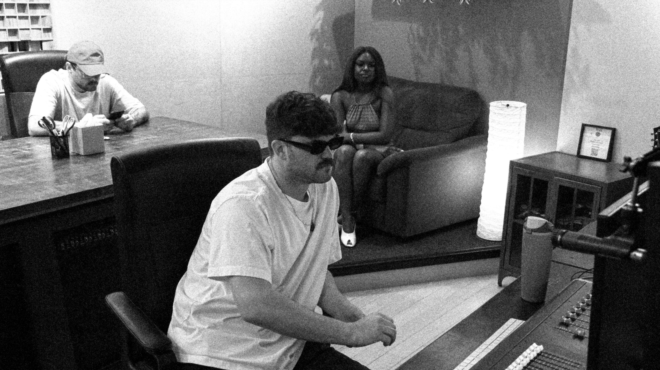 Three people in a recording studio, one sitting at a mixing console, two on a sofa, one wearing glasses and a white shirt.