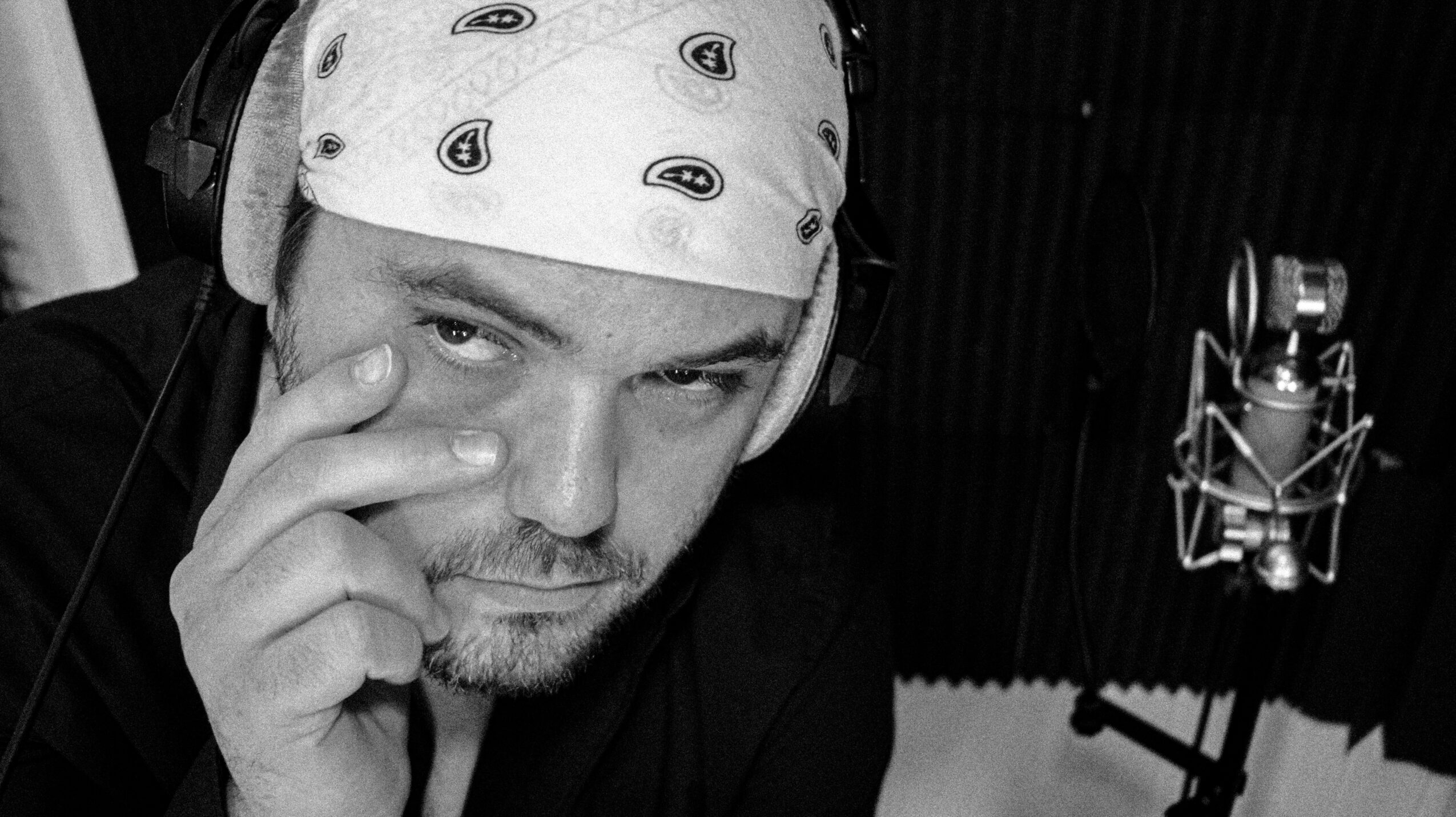 Man wearing a bandana and headphones, touching his face, in a recording studio with a microphone.