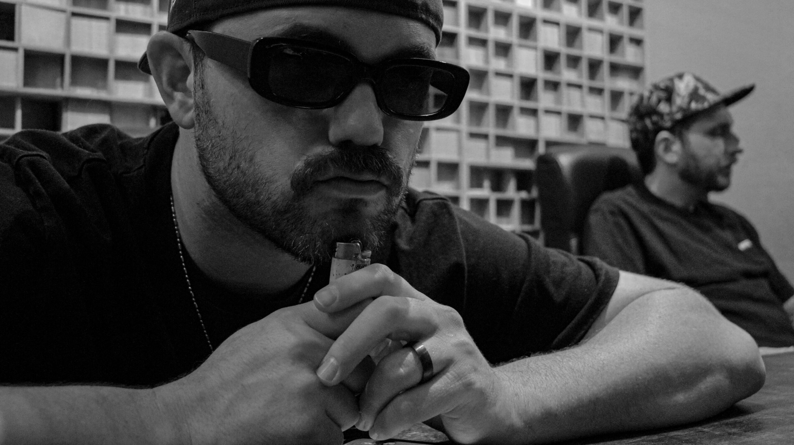 Two men sitting outdoors, one in foreground wearing sunglasses and holding a cigarette, the other in background wearing a cap.