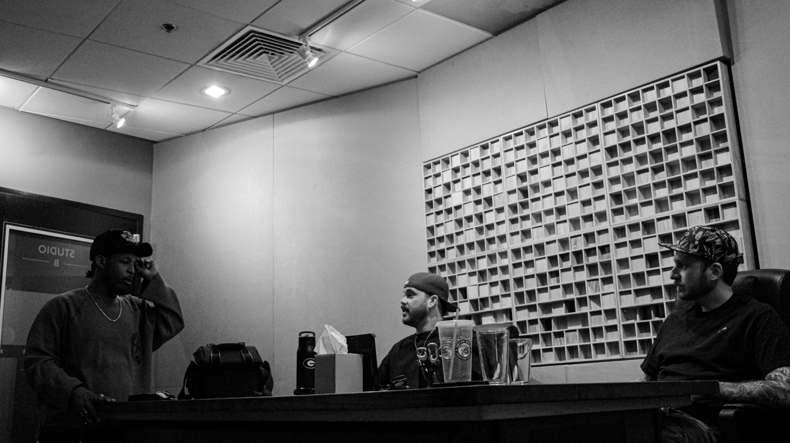 Three people in a room with a soundproof wall, two seated at a table and one standing, speaking into a microphone.