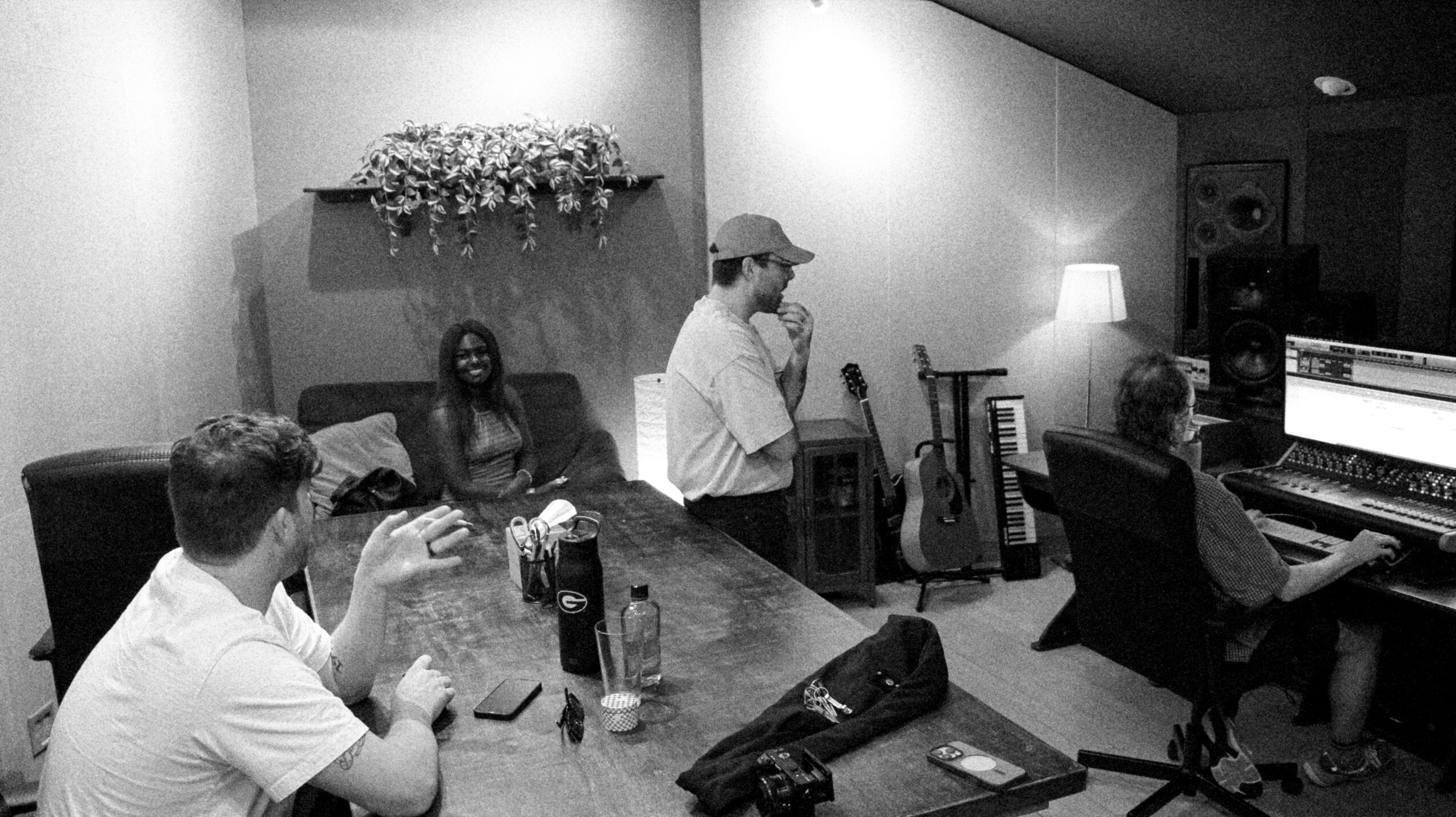 Four people in a music studio, one playing piano, others sitting or standing, with musical instruments and equipment visible.