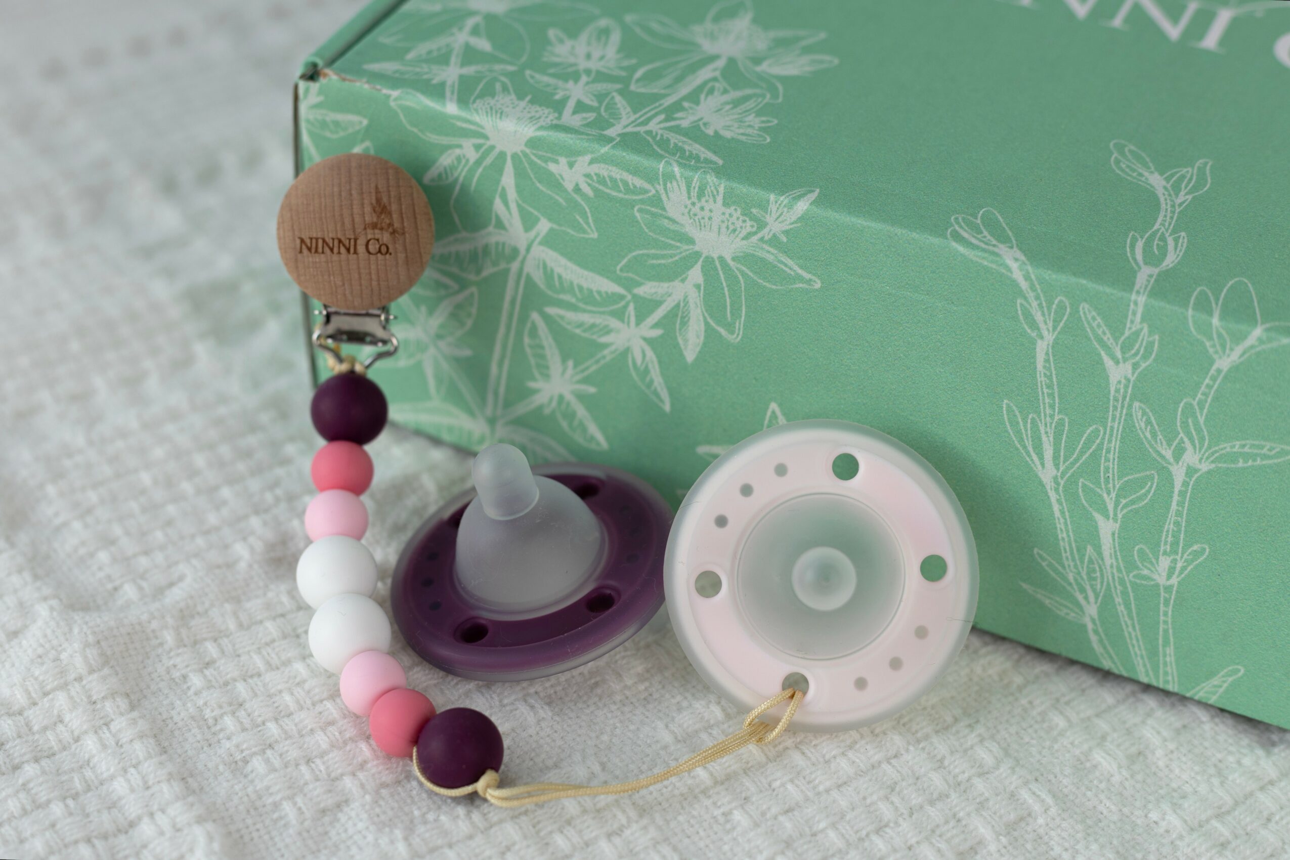 Baby pacifier and teething toy with beads in front of a green box, on a white textured surface.
