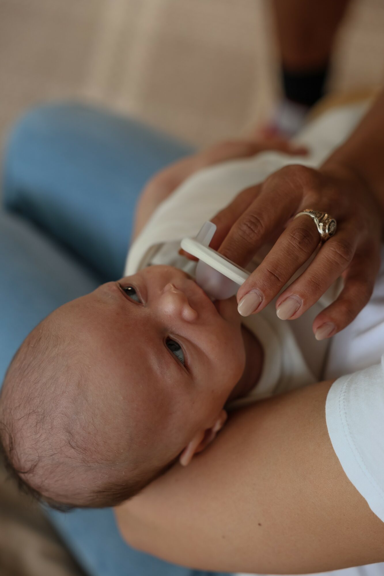 Baby lying on adult's arm, looking up, with adult holding a small object near baby's mouth, in a close-up view.