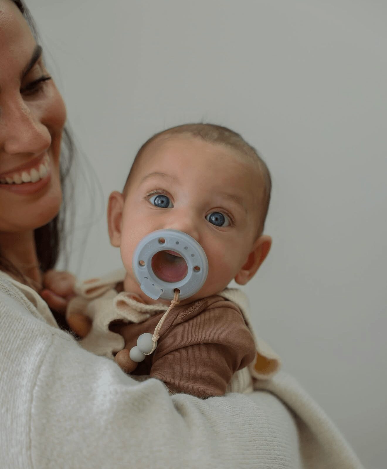 Baby with blue eyes and pacifier, held by a person, against a plain background.