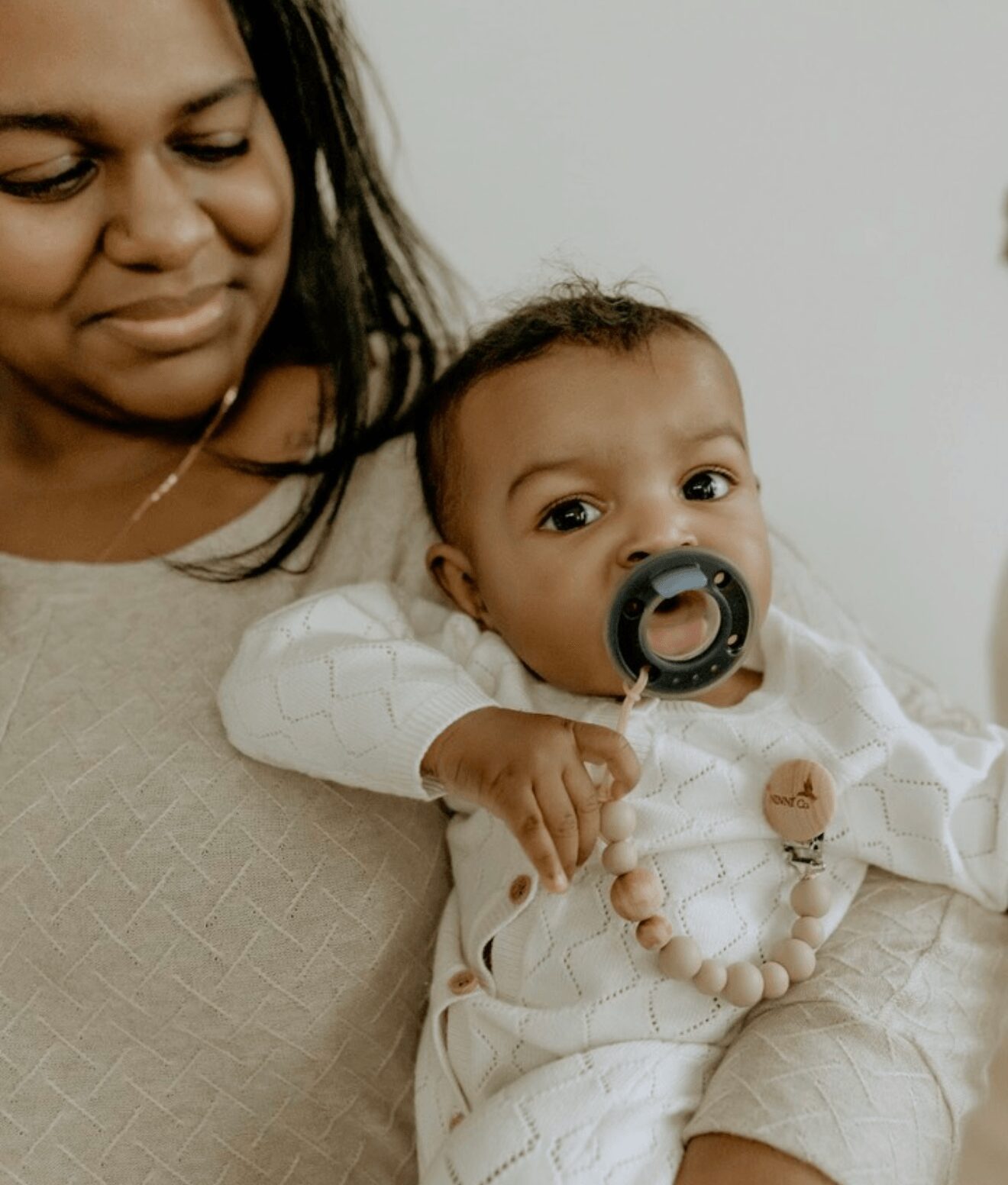 Adult holding a baby with a pacifier, both looking at the camera, neutral background, close-up shot.