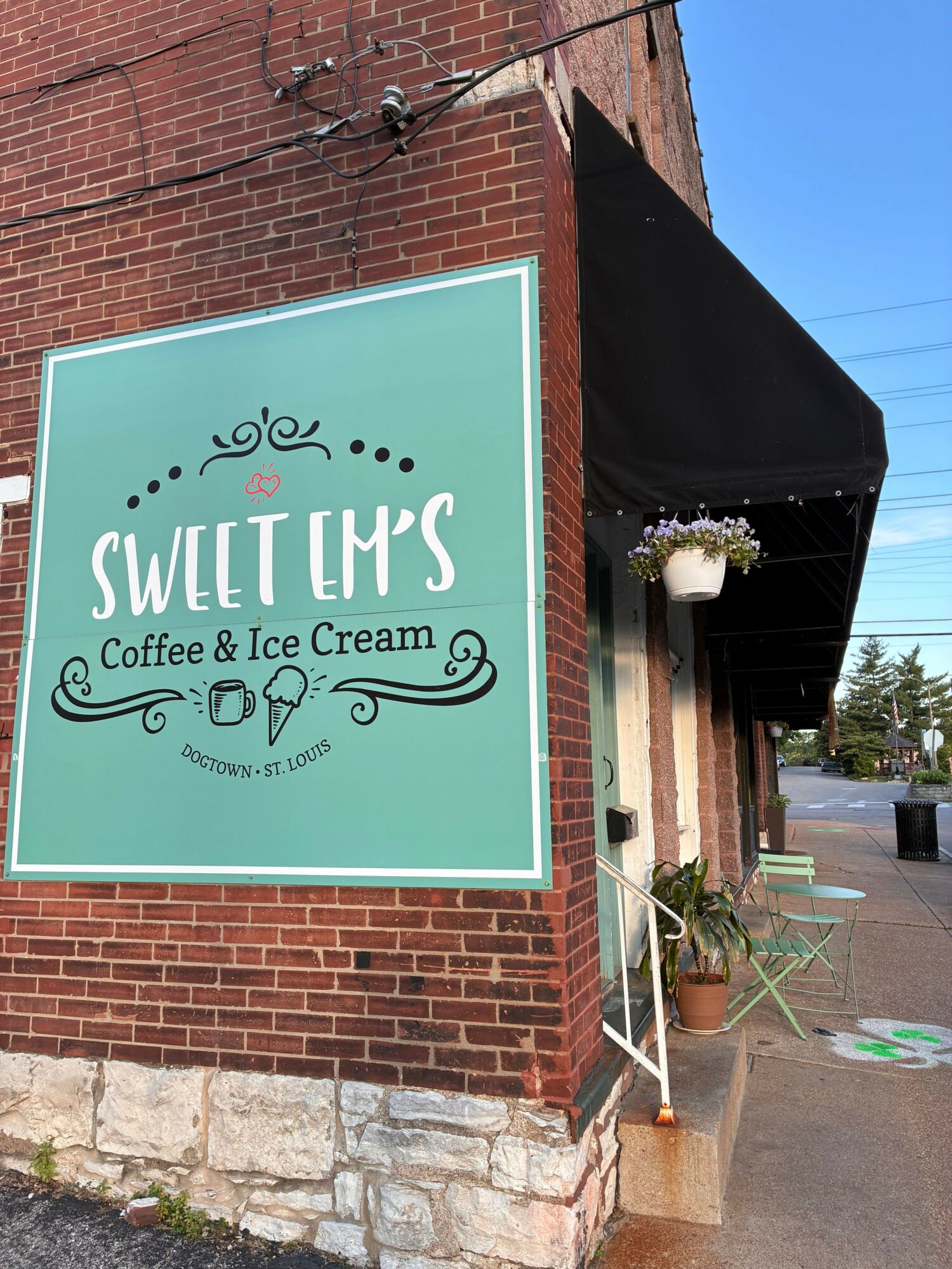 Sign for Sweet M's coffee and ice cream shop on brick wall, with outdoor seating and plants nearby.
