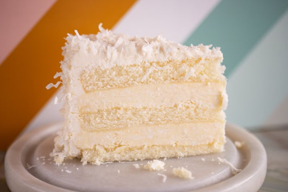 Slice of layered white cake with coconut frosting on a plate, with colorful geometric background