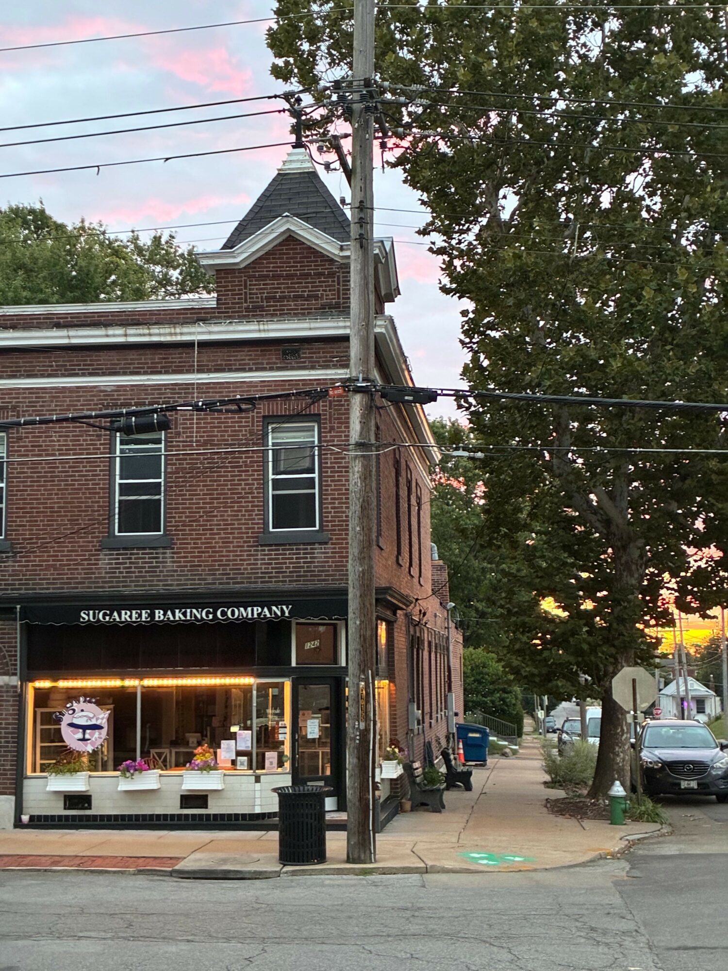 Brick building with a sign reading 'SCHOOL OF THE ARTS' and a person sitting outside, trees and parked cars nearby.