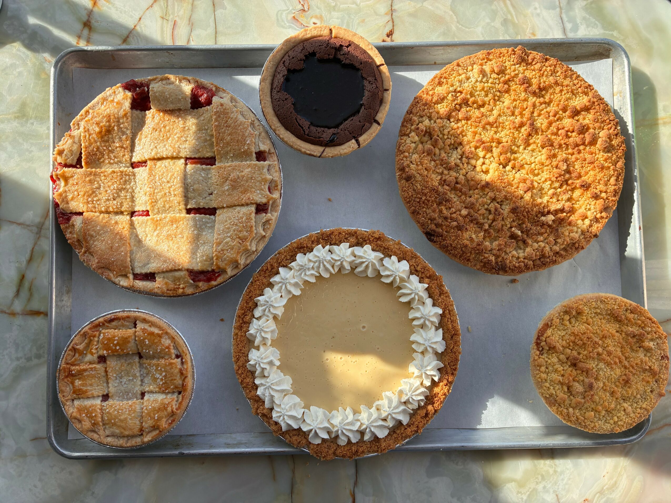 Assorted baked goods on a tray, including a pie, muffin, cookie, and cake with white frosting, on a marble surface.
