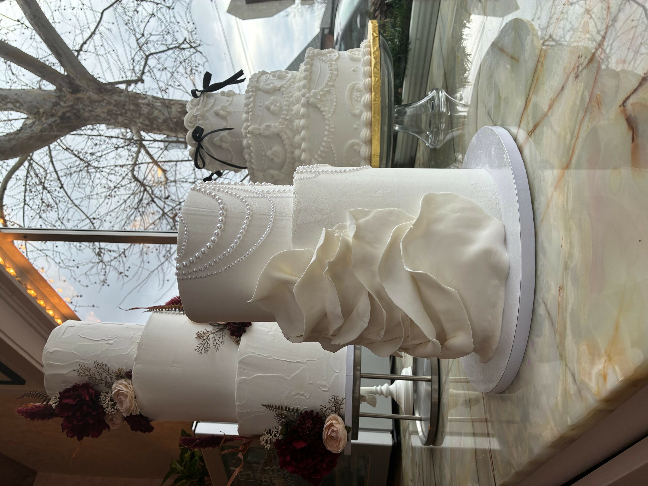 Three white cakes with decorative icing and flowers on a marble surface outdoors, with trees and a building in background.