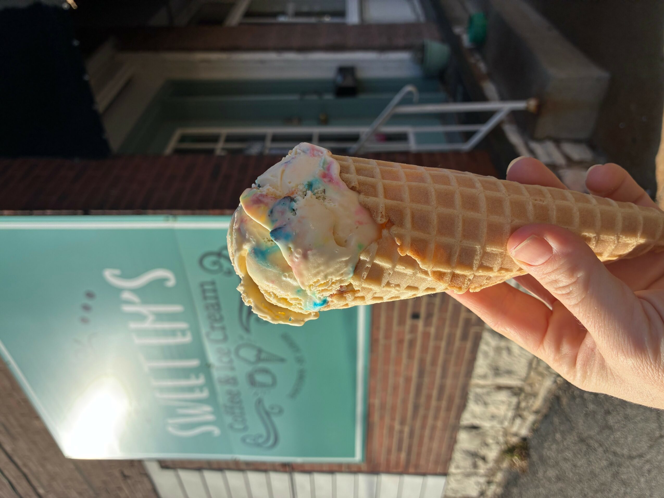 Hand holding a cone with colorful ice cream, background shows a sign and building exterior.