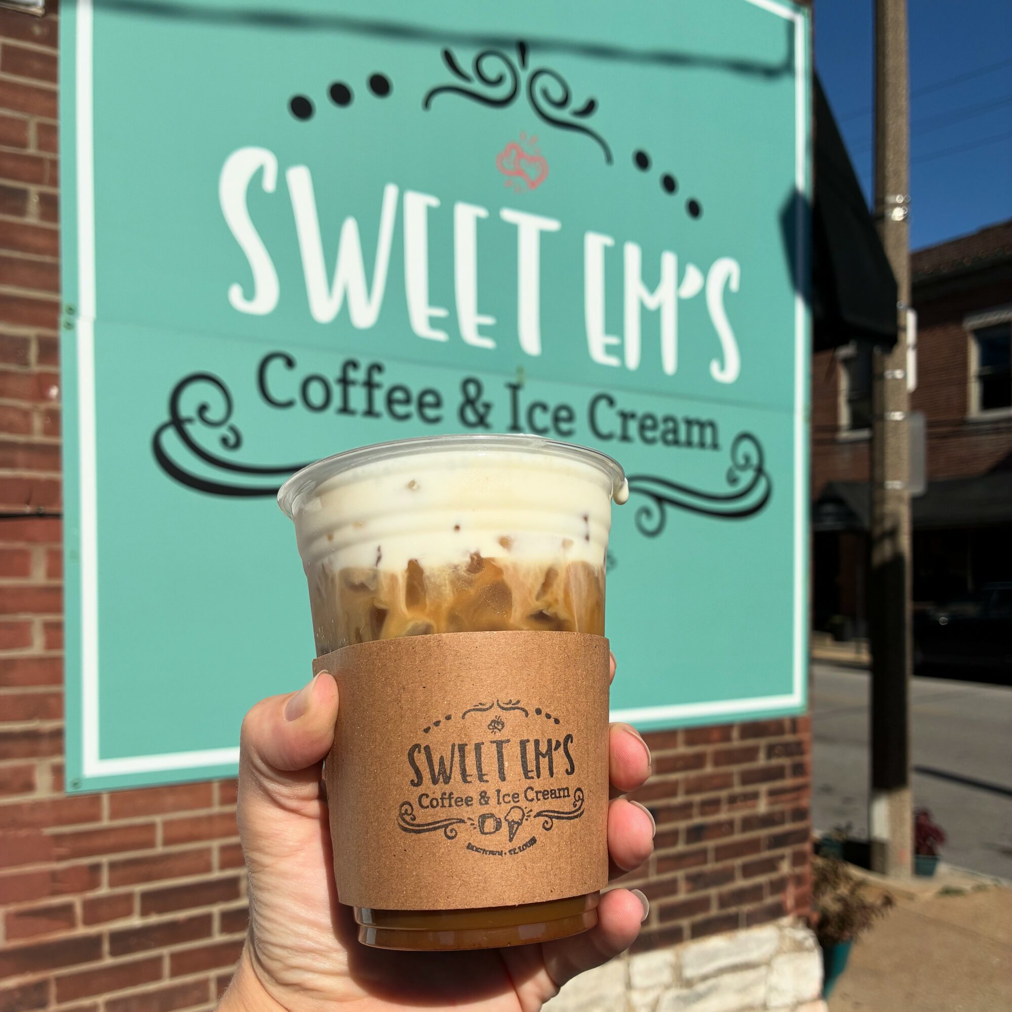 Hand holding iced coffee with a brown sleeve in front of a teal sign for Sweet Lips Coffee & Ice Cream, brick building background.