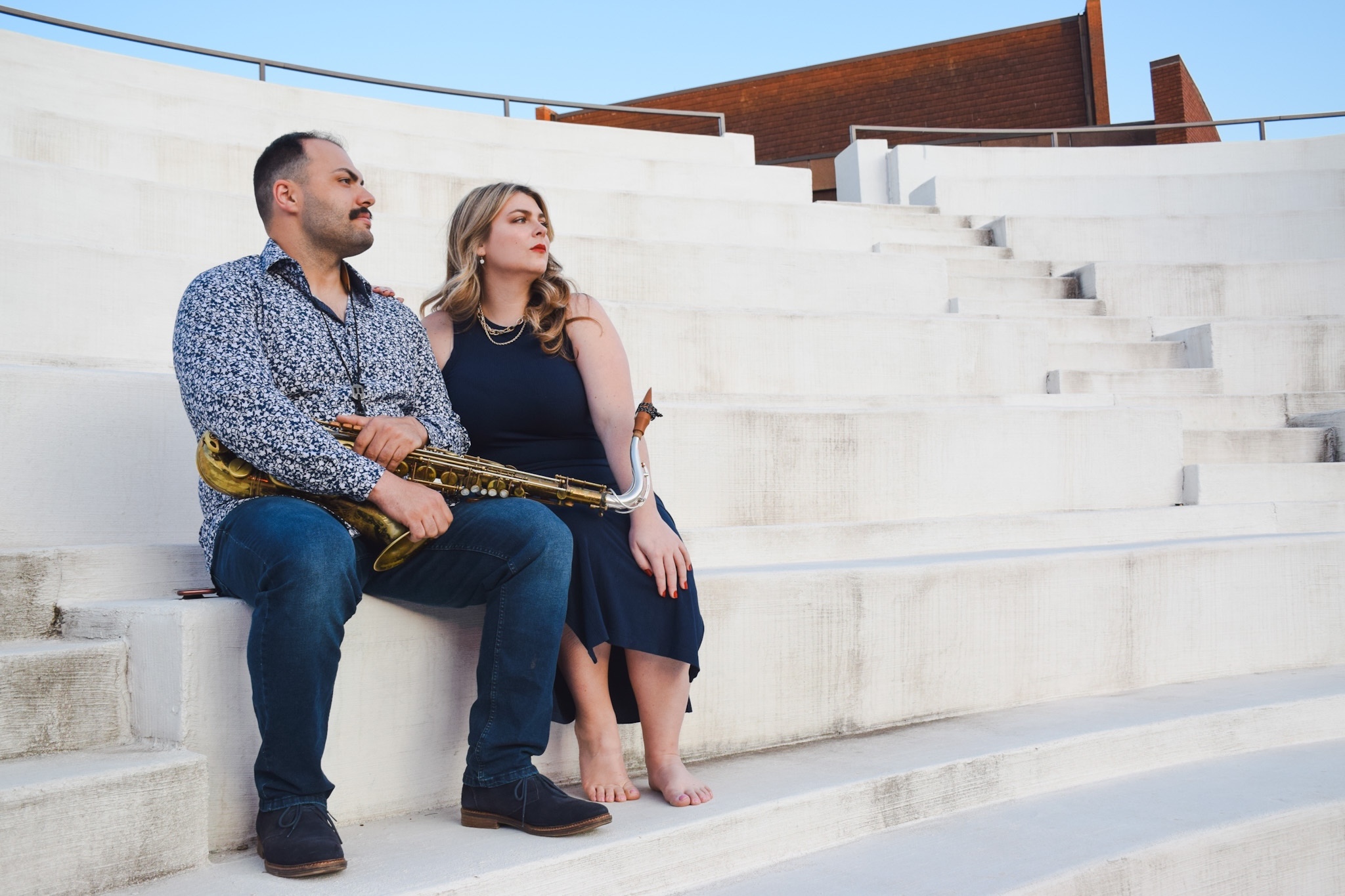 Two people sitting on concrete steps, one holding a saxophone, outdoors with modern buildings in background.