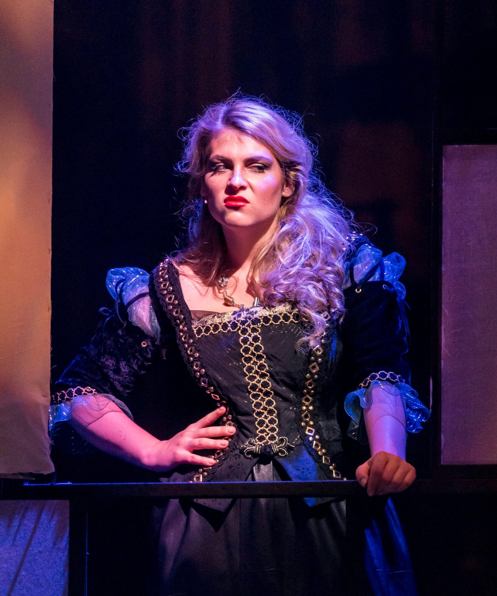 Woman with long, wavy blonde hair in a dark, ornate dress standing on stage with a serious expression.