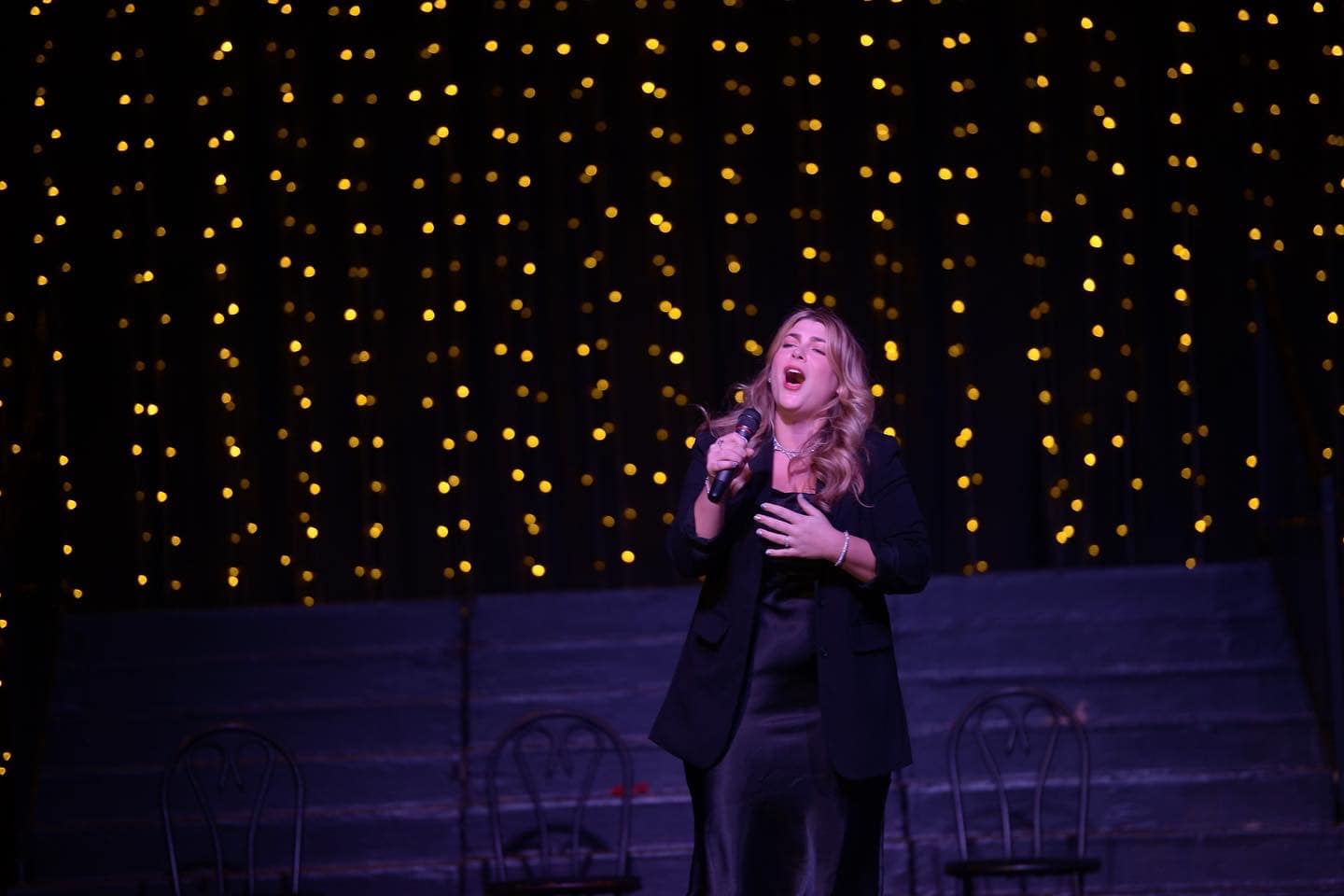 A woman singing on stage with a black background and yellow string lights, holding a microphone, with chairs behind her.