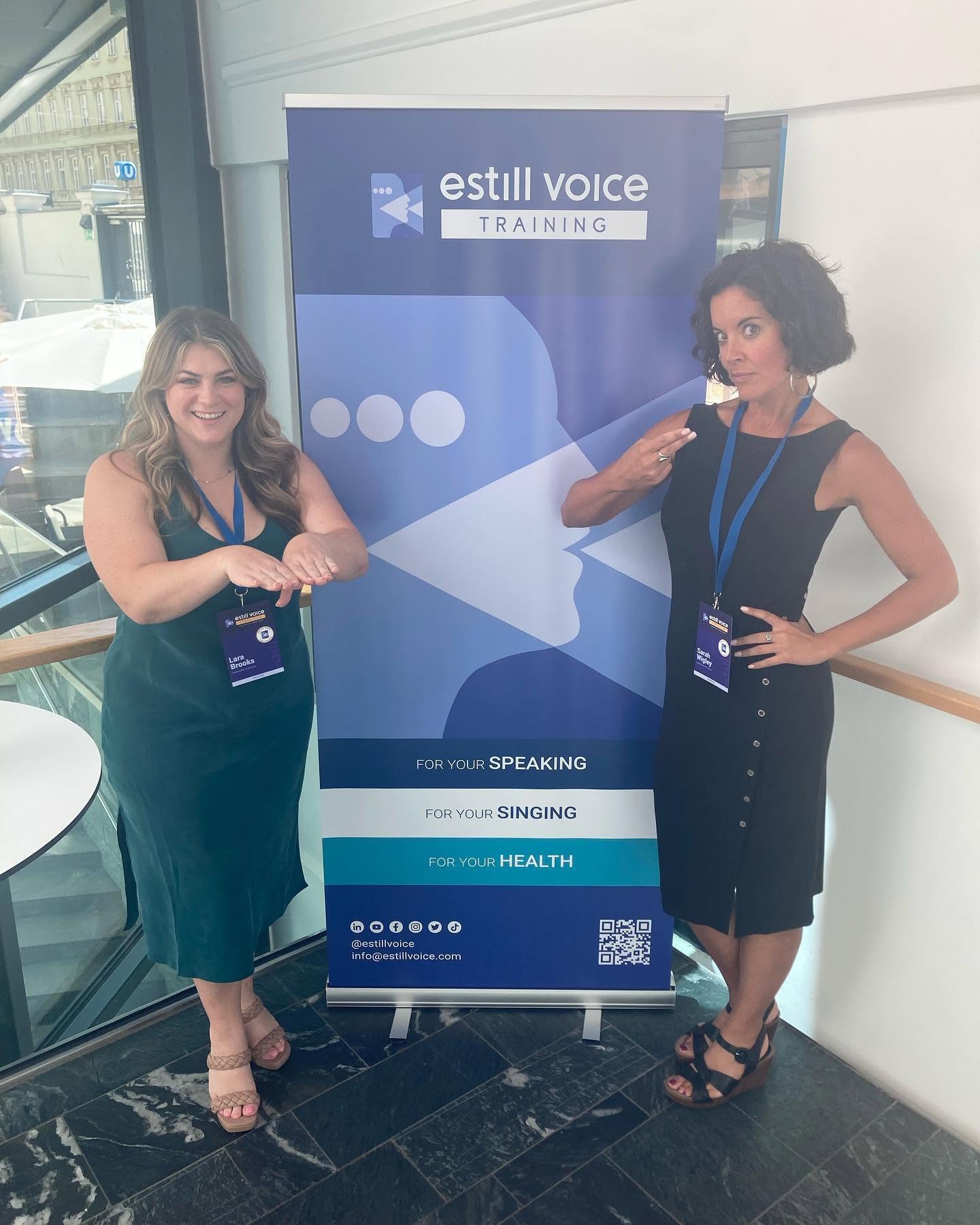 Two women stand next to a blue and white banner for Estill Voice Training, posing indoors with one pointing at the other.