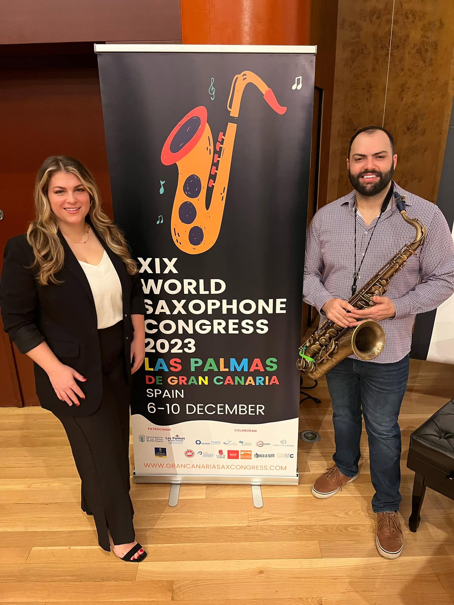 Two people, a woman and a man holding a saxophone, stand next to a banner for the 2023 World Saxophone Congress in Las Palmas, Spain.
