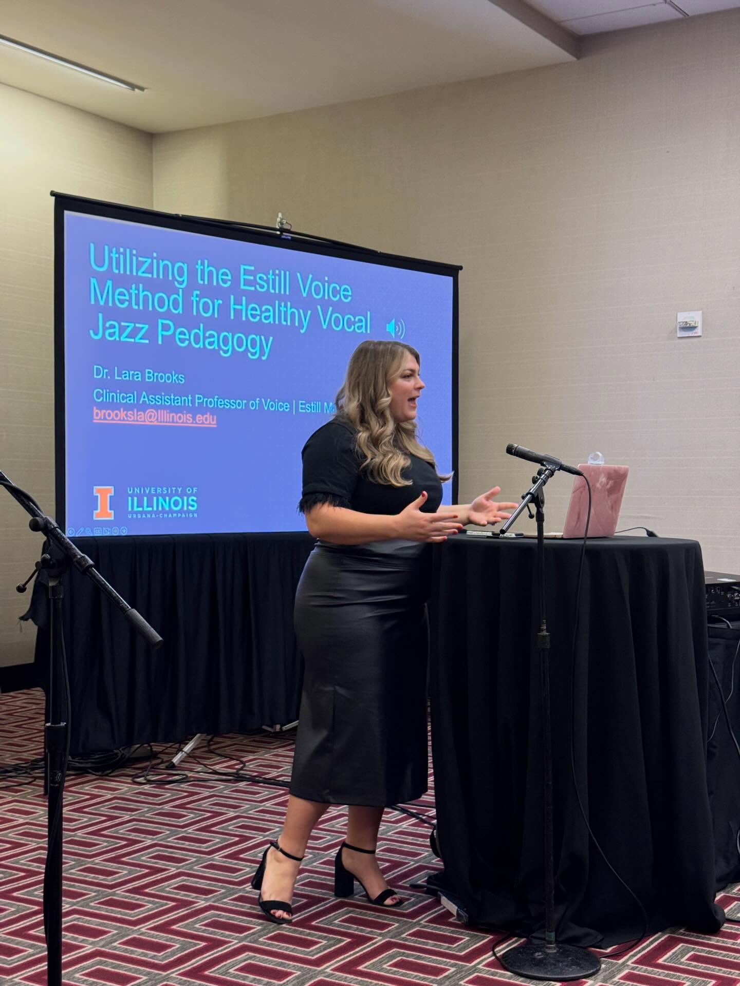 Woman standing at a podium giving a presentation in a conference room, with a screen displaying a slide titled 'Utilizing the Estill Voice Method for Healthy Vocal & Jazz Pedagogy.'