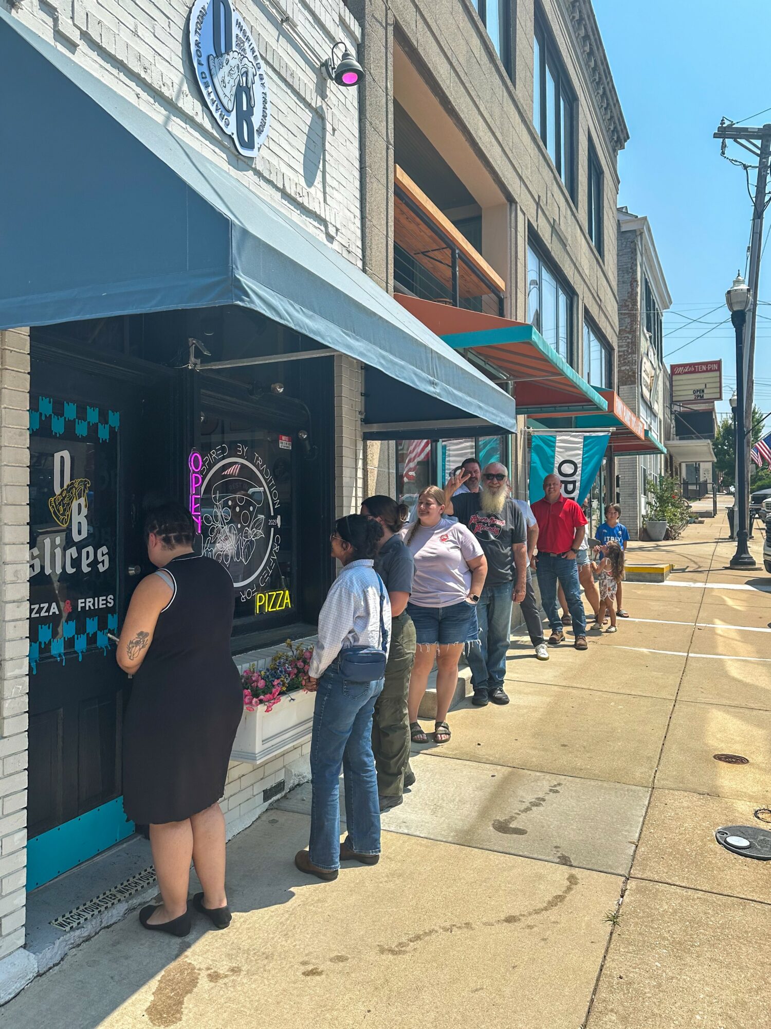 People standing in line outside a pizza shop on a city sidewalk, with buildings and a street visible.