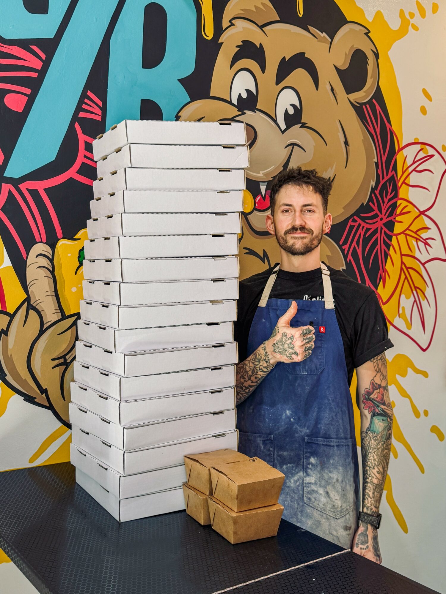 Man with tattoos and beard in apron stands next to a tall stack of pizza boxes and three smaller boxes, colorful mural background.