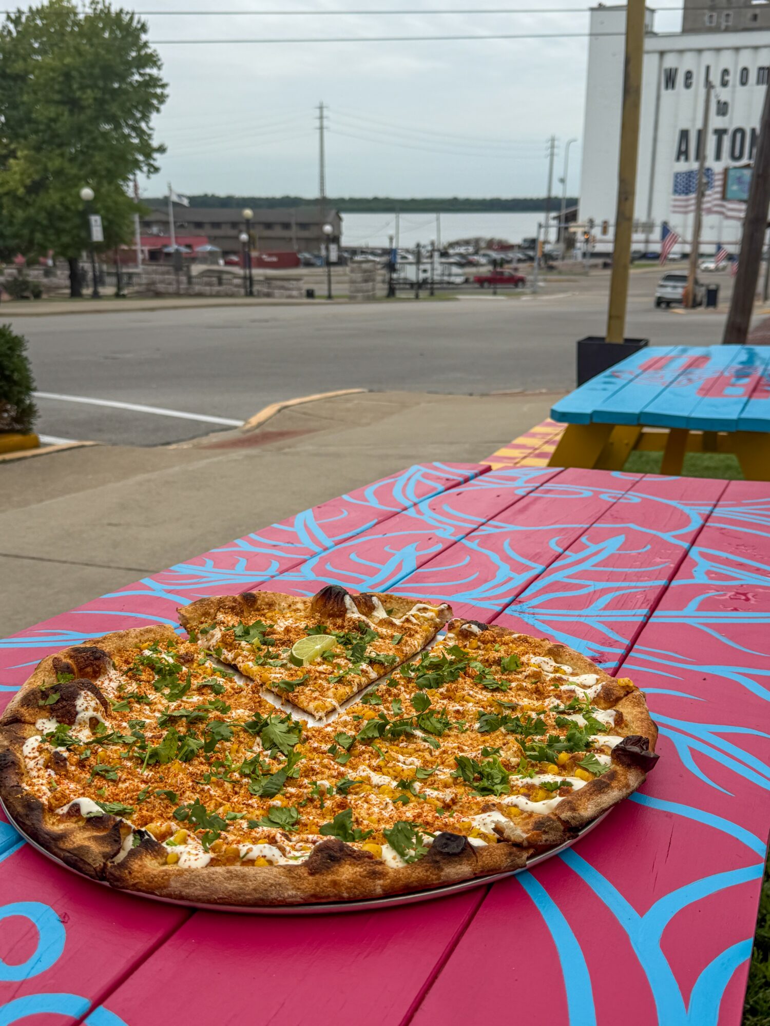 Large pizza with toppings on a colorful table outside, with a street and buildings in the background.