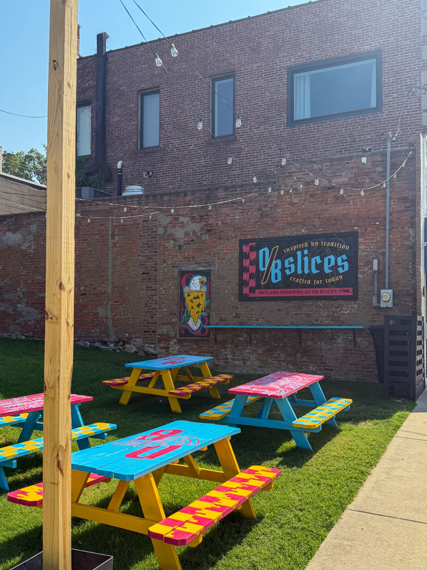 Outdoor seating area with colorful picnic tables in front of a brick building, sign reads 'Slices' with a pizza graphic.