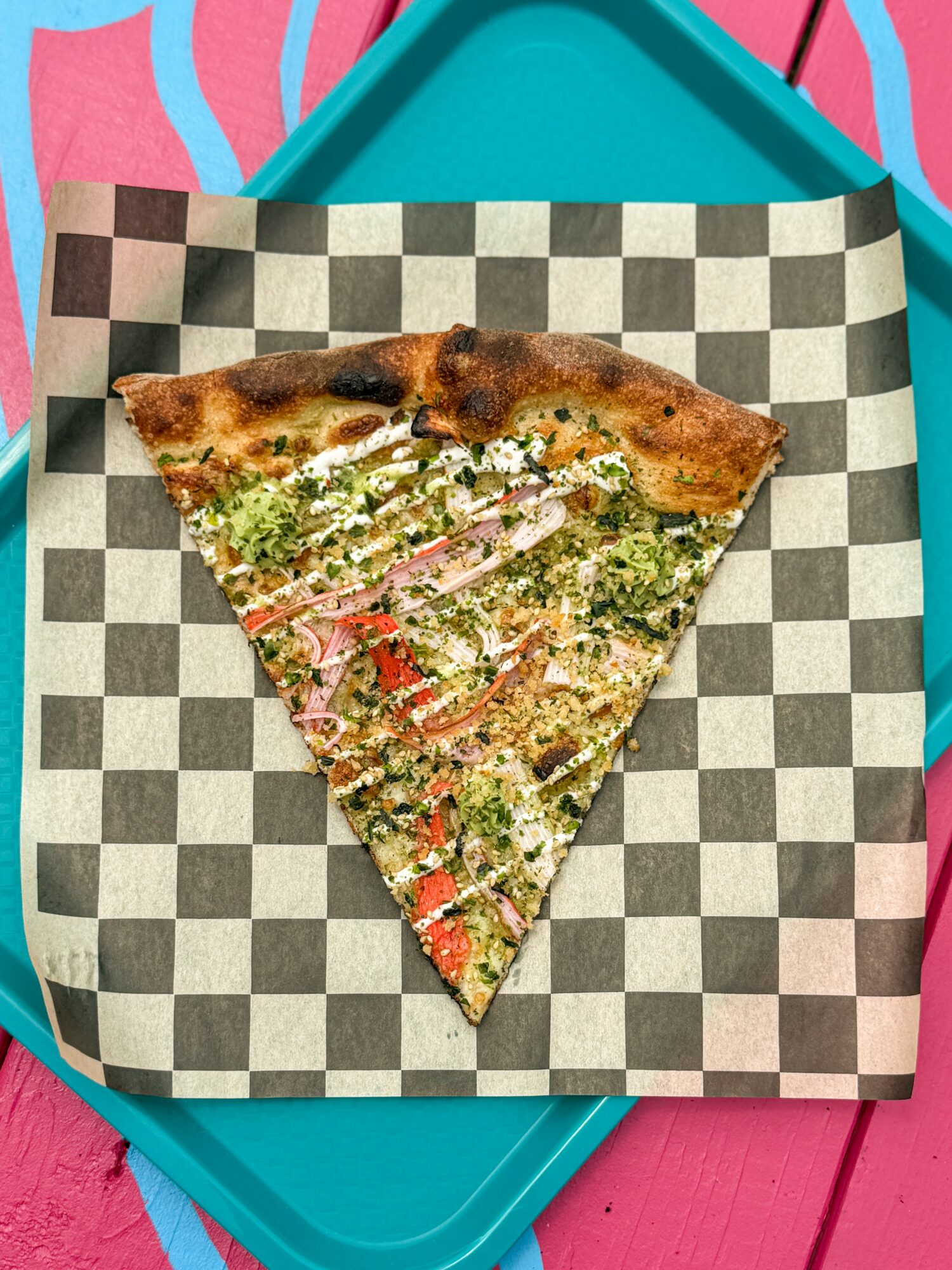 A slice of pizza on checkered paper, placed on a teal tray with a pink background.