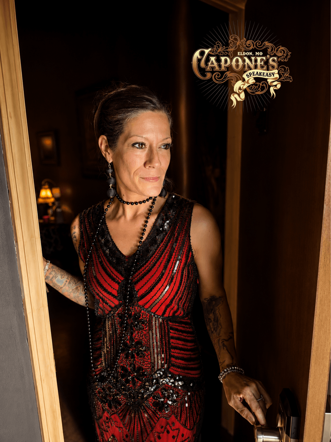 Woman in a red and black dress with lace sleeves, standing in a doorway, looking to the side, with a logo in the top right corner.