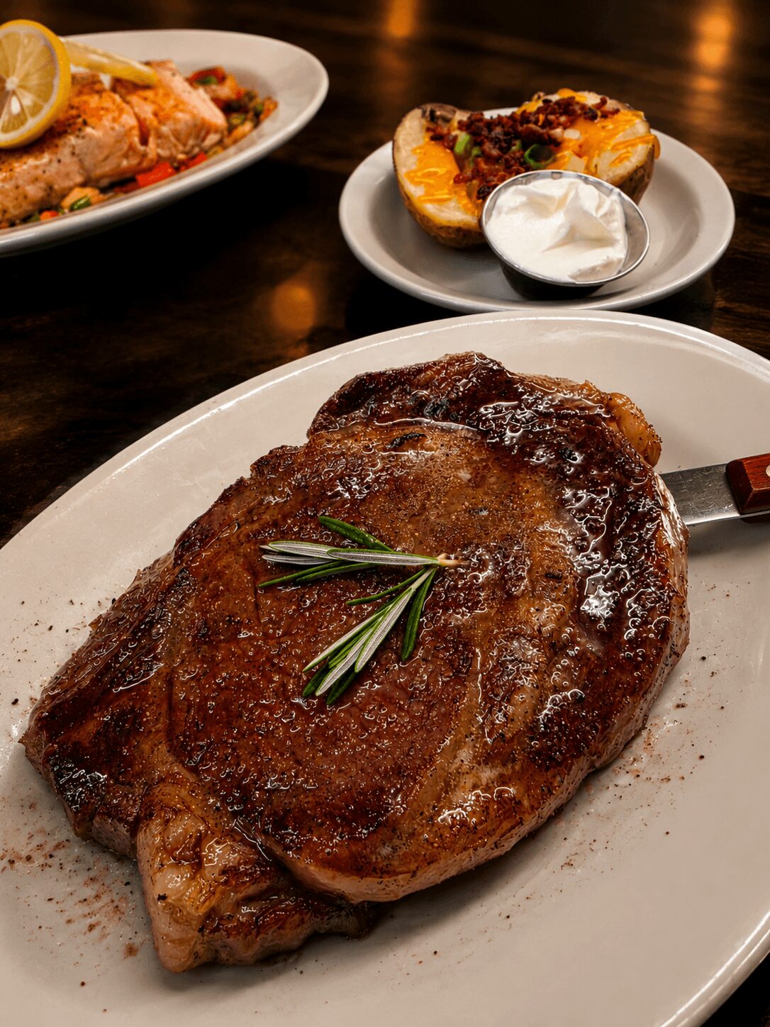 A cooked steak garnished with rosemary on a white plate, with side dishes in the background.