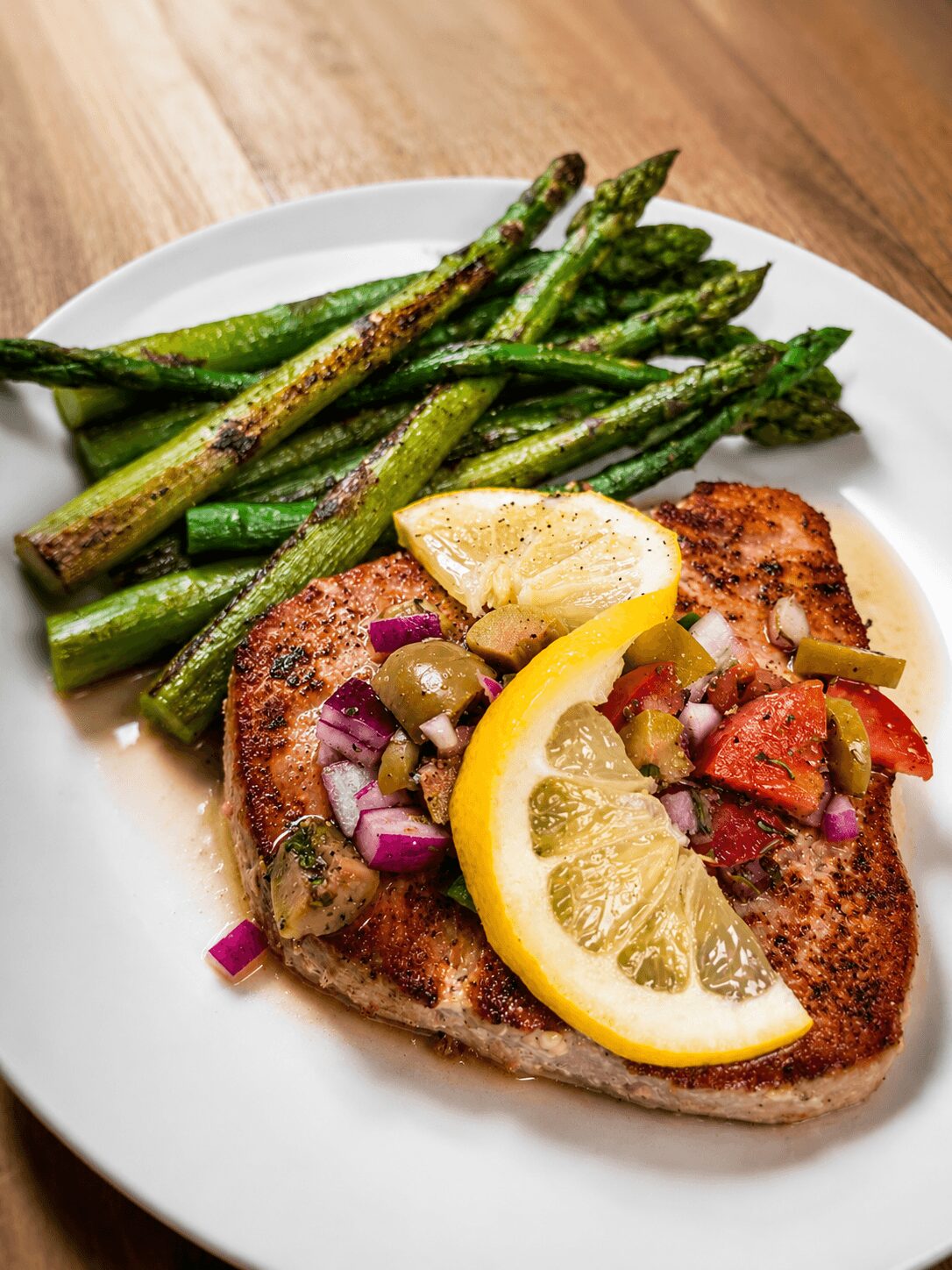 Grilled fish fillet with lemon wedge, topped with chopped vegetables, served with roasted asparagus on a white plate.
