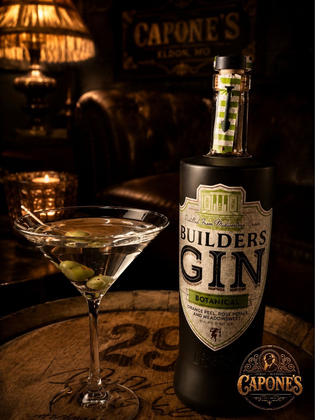 A bottle of Builders Gin and a martini glass with an olive inside on a wooden surface, dimly lit background.