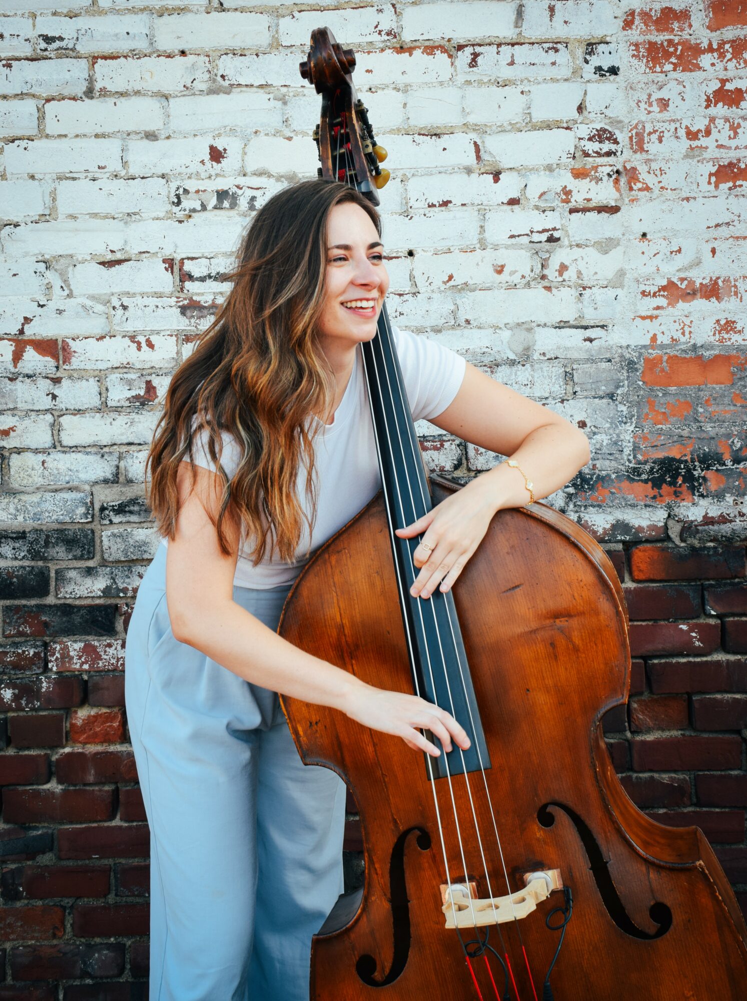 Woman with long hair smiling, holding a double bass against a brick wall.
