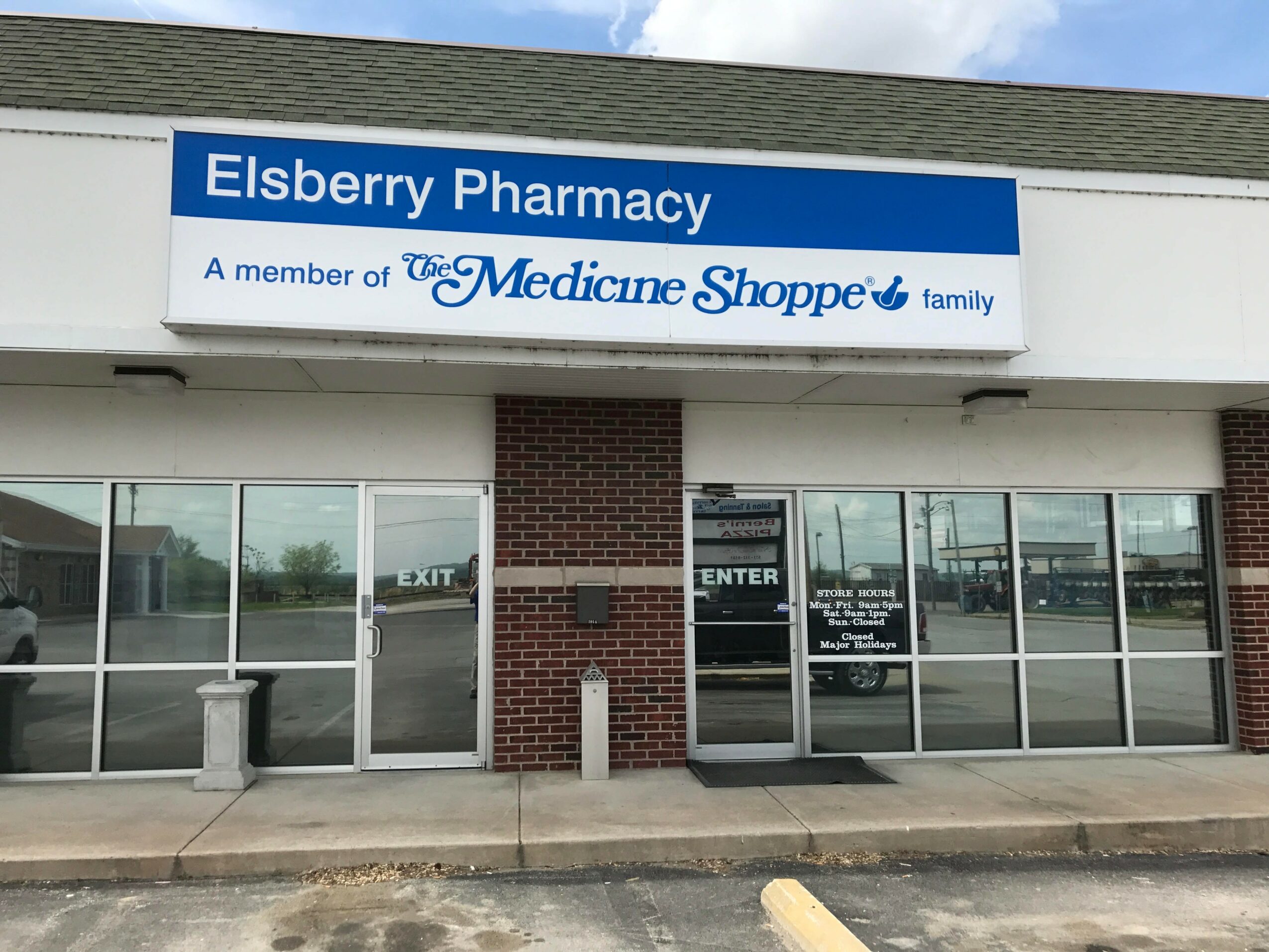 Storefront with sign reading 'Elsberry Pharmacy' and 'A member of the Medicine Shoppe family' above entrance, glass windows, and brick column.