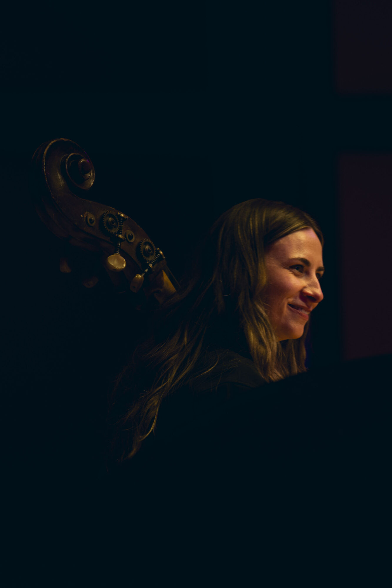Woman with long hair smiling, dark background, musical instrument partially visible, side profile.