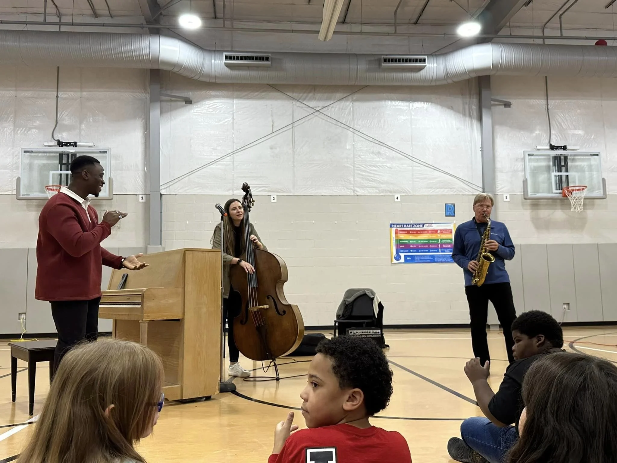 Three musicians perform in a gymnasium with basketball hoops, audience seated on the floor, and a colorful chart on the wall.