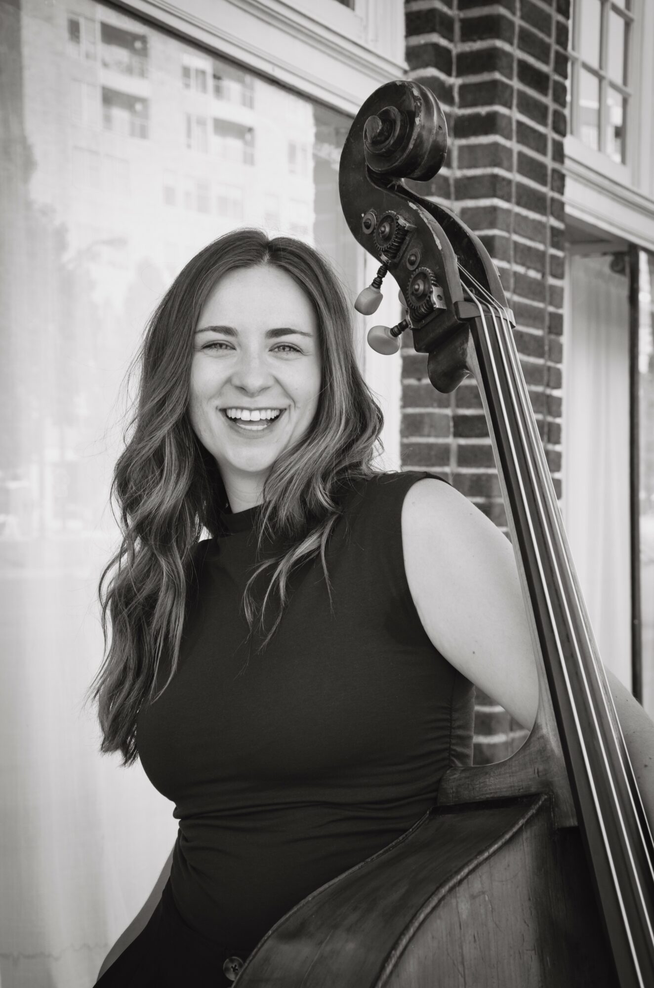 Woman smiling with a double bass, standing outside a building with large windows and brick wall, in black and white.