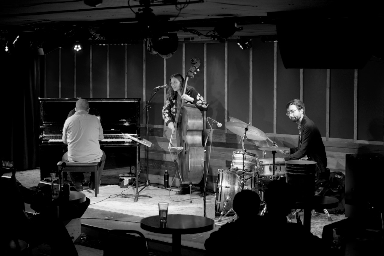 Three musicians perform on stage: a pianist, a double bassist, and a drummer, with audience silhouettes in foreground.